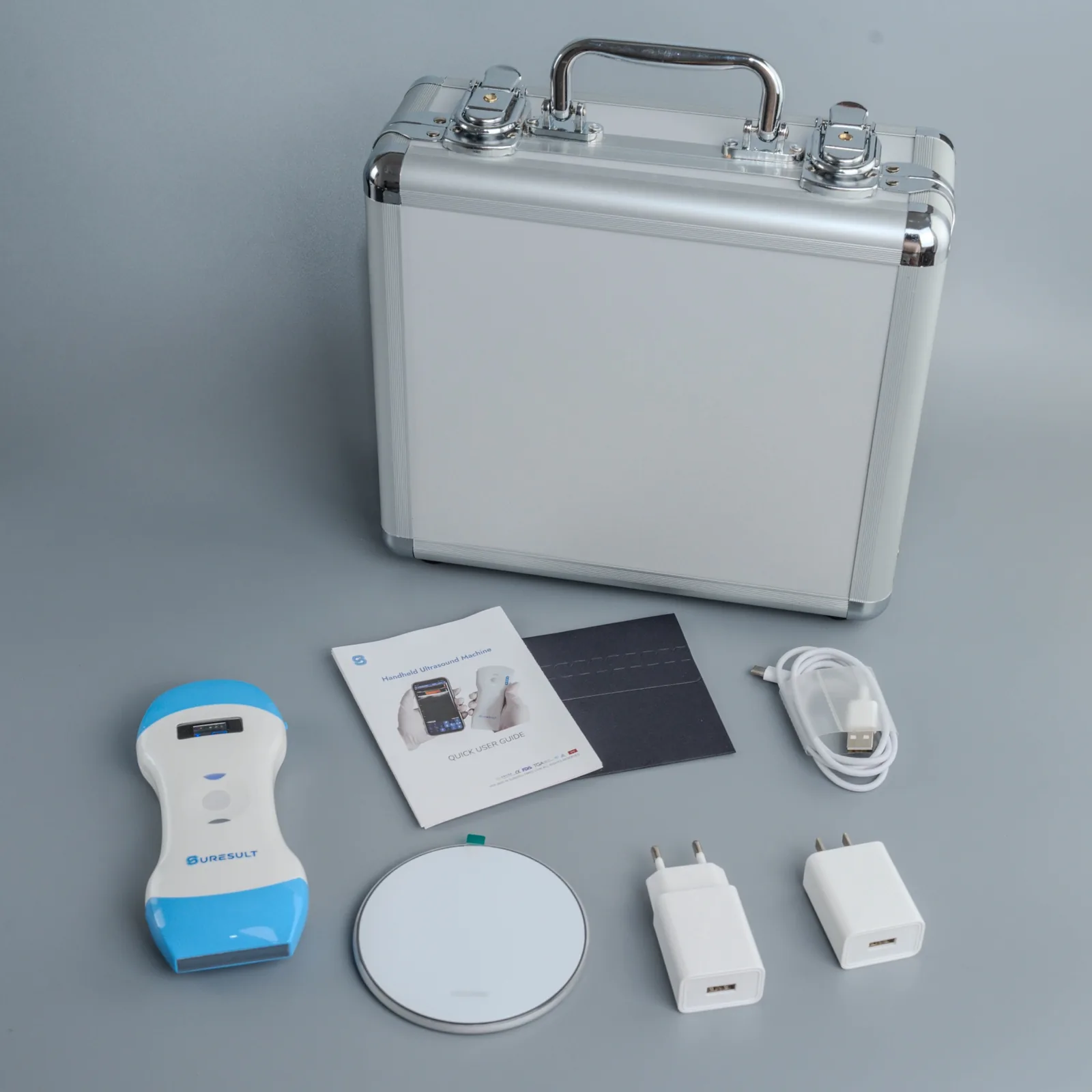 The Suresult aluminium case behind a handheld probe with a blue tip, the quick user guide, a wireless charging pad, two plugs and a charging cable, laid out on a grey studio ground